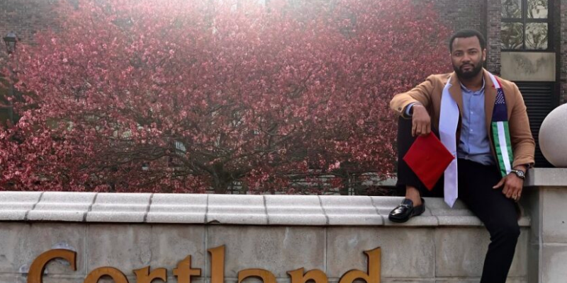 A student posing outside the Cortland sign wearing their grad sash.